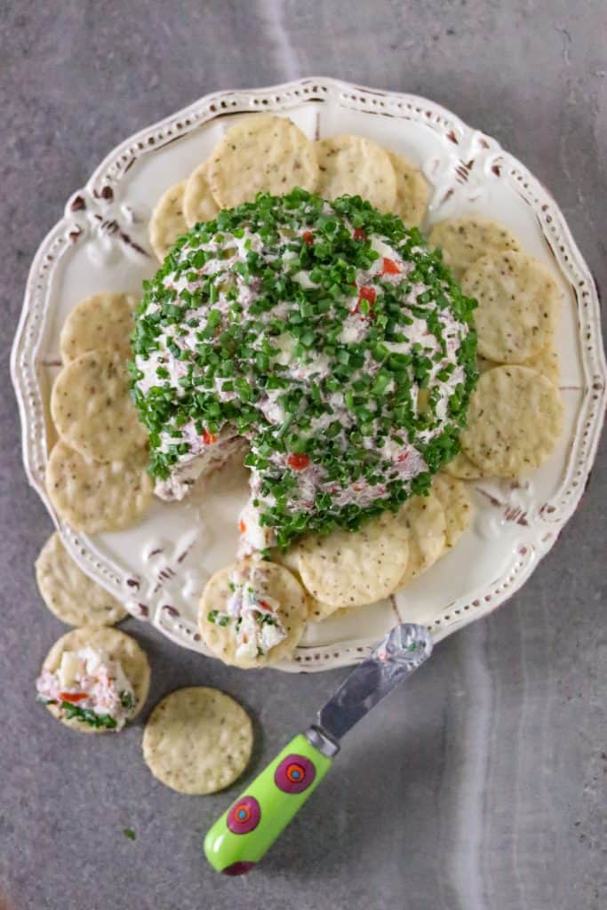 A cheese ball covered with chopped chives on a plate with crackers for Mardi Gras Food Recipes.
