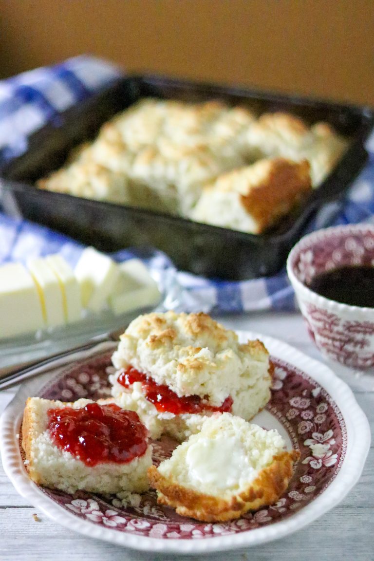 A pan of biscuits with a buttered biscuit with jam on a plate next to it with a cup of coffee.
