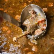 A pot of goose gumbo with a ladle full of gumbo juice, a goose thigh quarter, and sliced sausage.