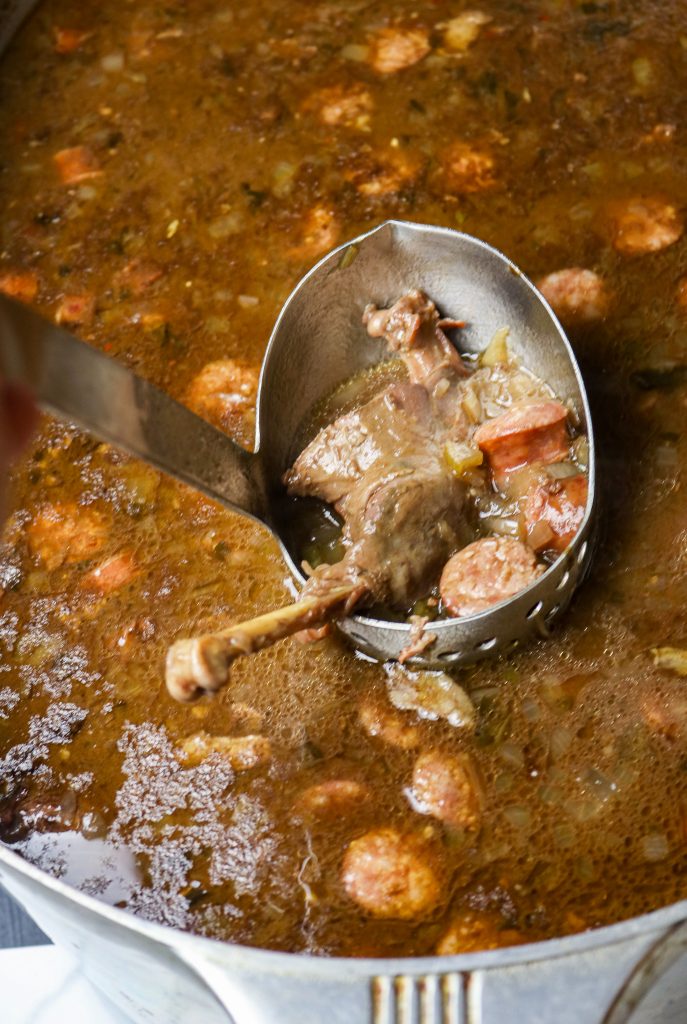 A pot of goose gumbo with a ladle full of gumbo juice, a goose thigh quarter, and sliced sausage.