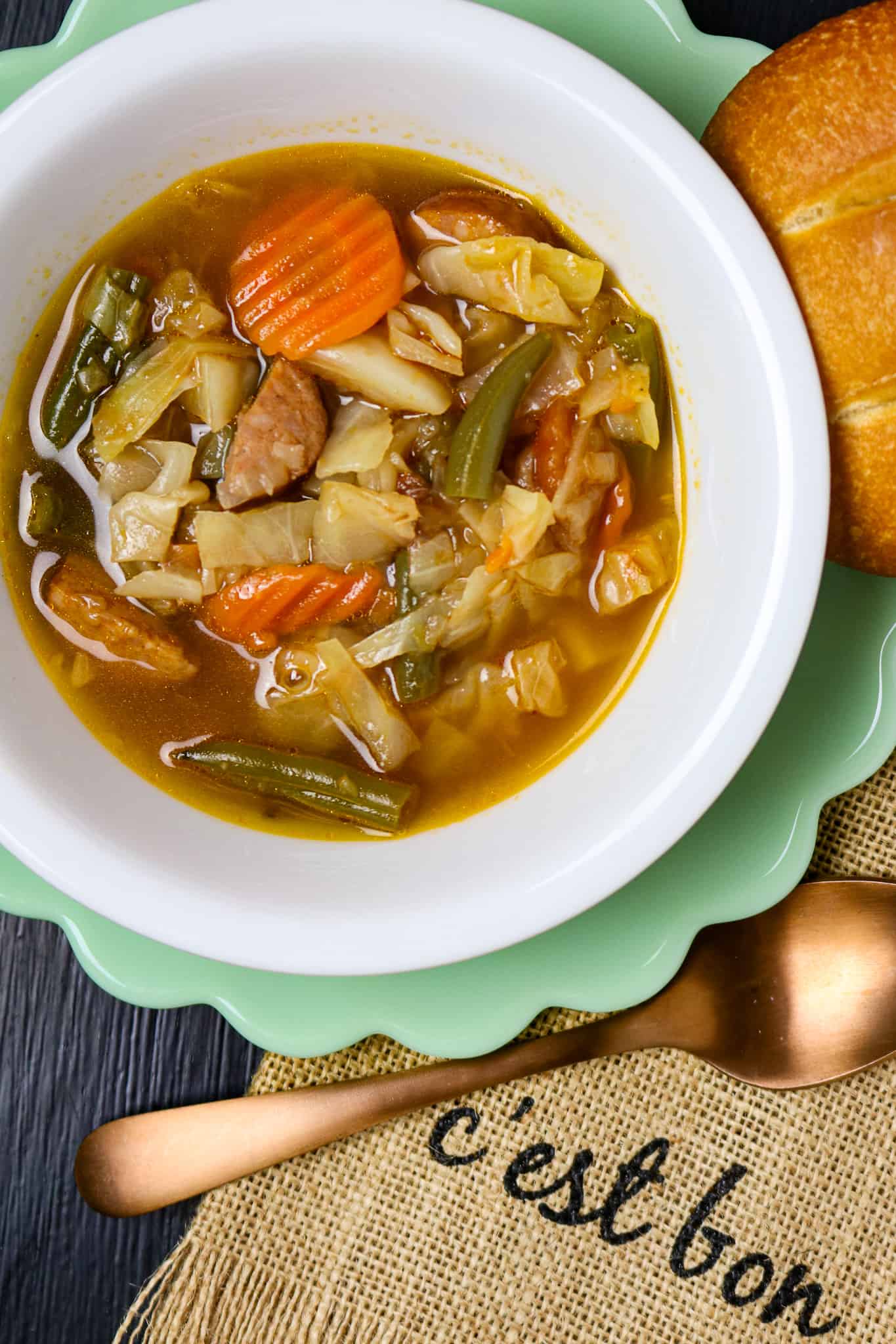 A bowl of cabbage soup with andouille sausage with a napkin that says cest' bon and a spoon.