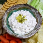 A bowl of pickle dip surrounded by carrots, celery, chip, and crackers.