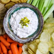 A bowl of pickle dip surrounded by carrots, celery, chip, and crackers.
