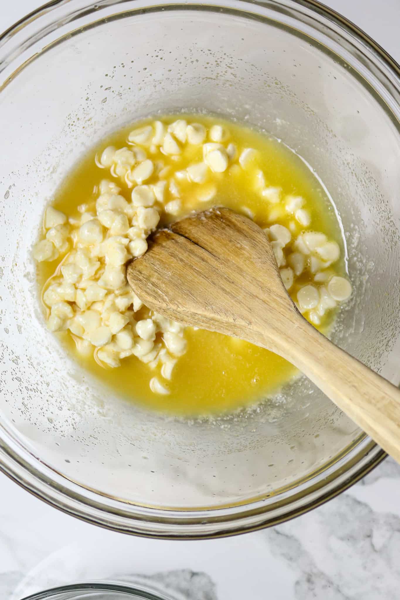 A wooden spoon stirring butter and white chocolate in a glass bowl.