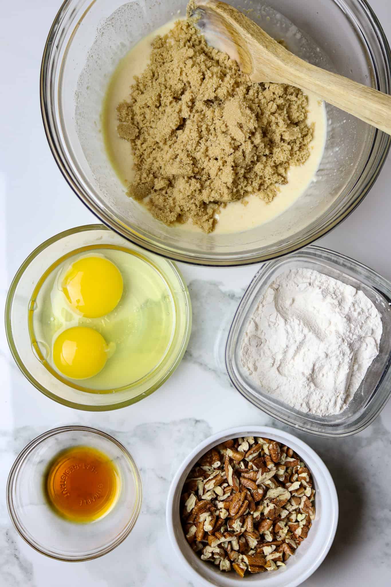 A glass bowl of brown sugar next to a bowl of eggs, flour, pecans, and vanilla.
