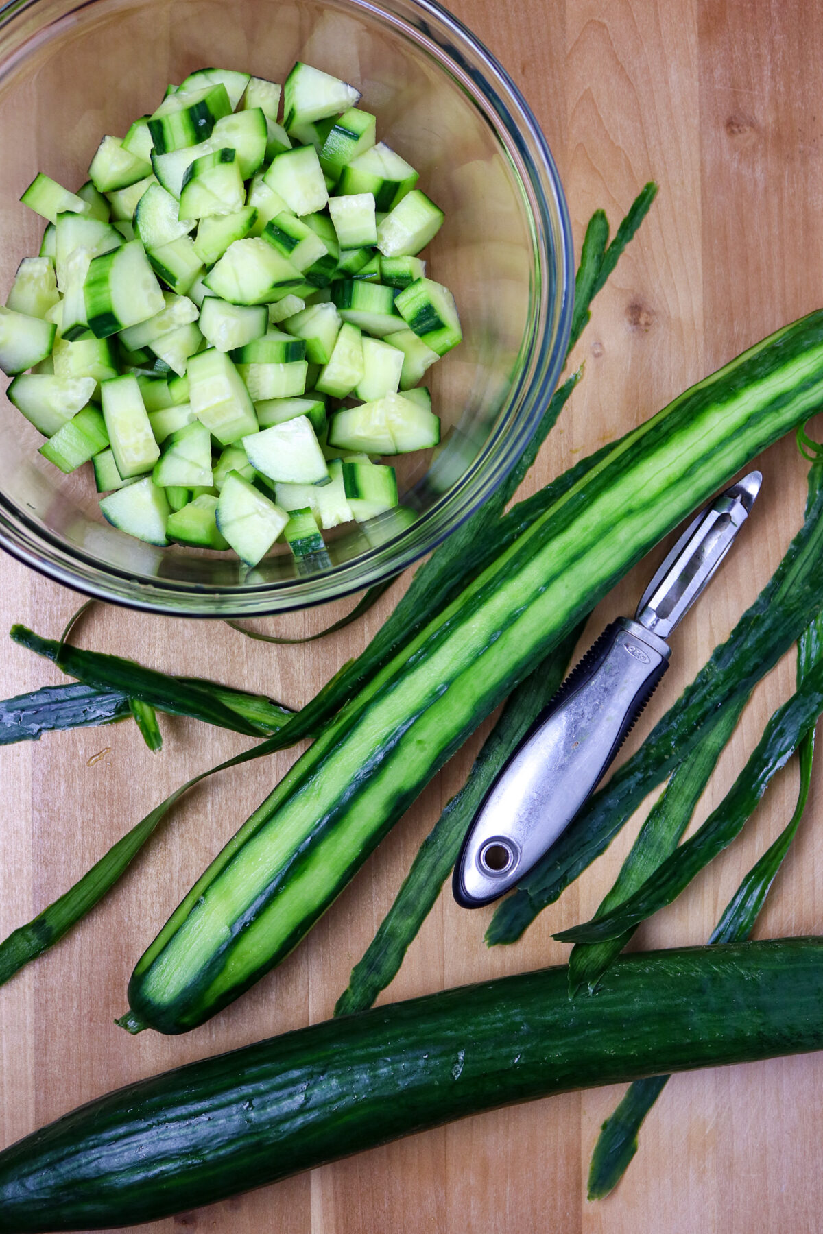 A bowl of sliced cucumber, a vegetable peeler, and an English cucumber on a wooden board.