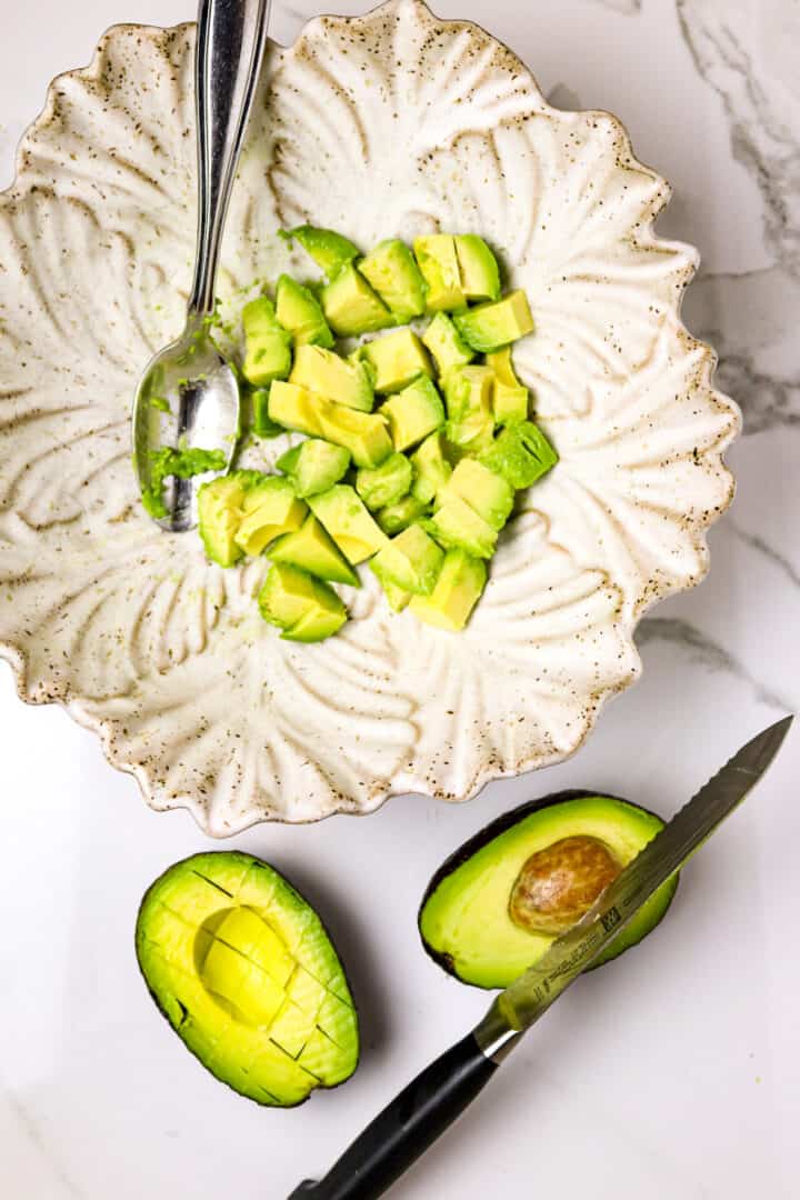 Cubed avocado in a white bowl with a silver spoon and a cut avocado beside with a knife stuck in the pit.