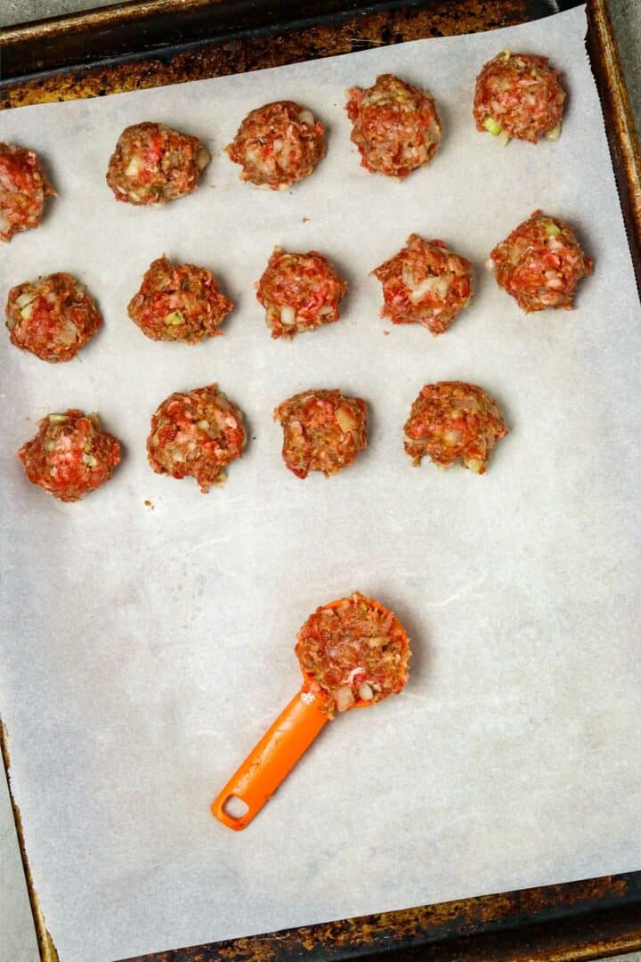 Small meatballs lined on a paper-lined cookie sheet with a tablespoon full in maiddle of pan.