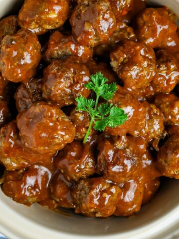 A bowl of party meatballs in sauce garnished with parsley leaves.