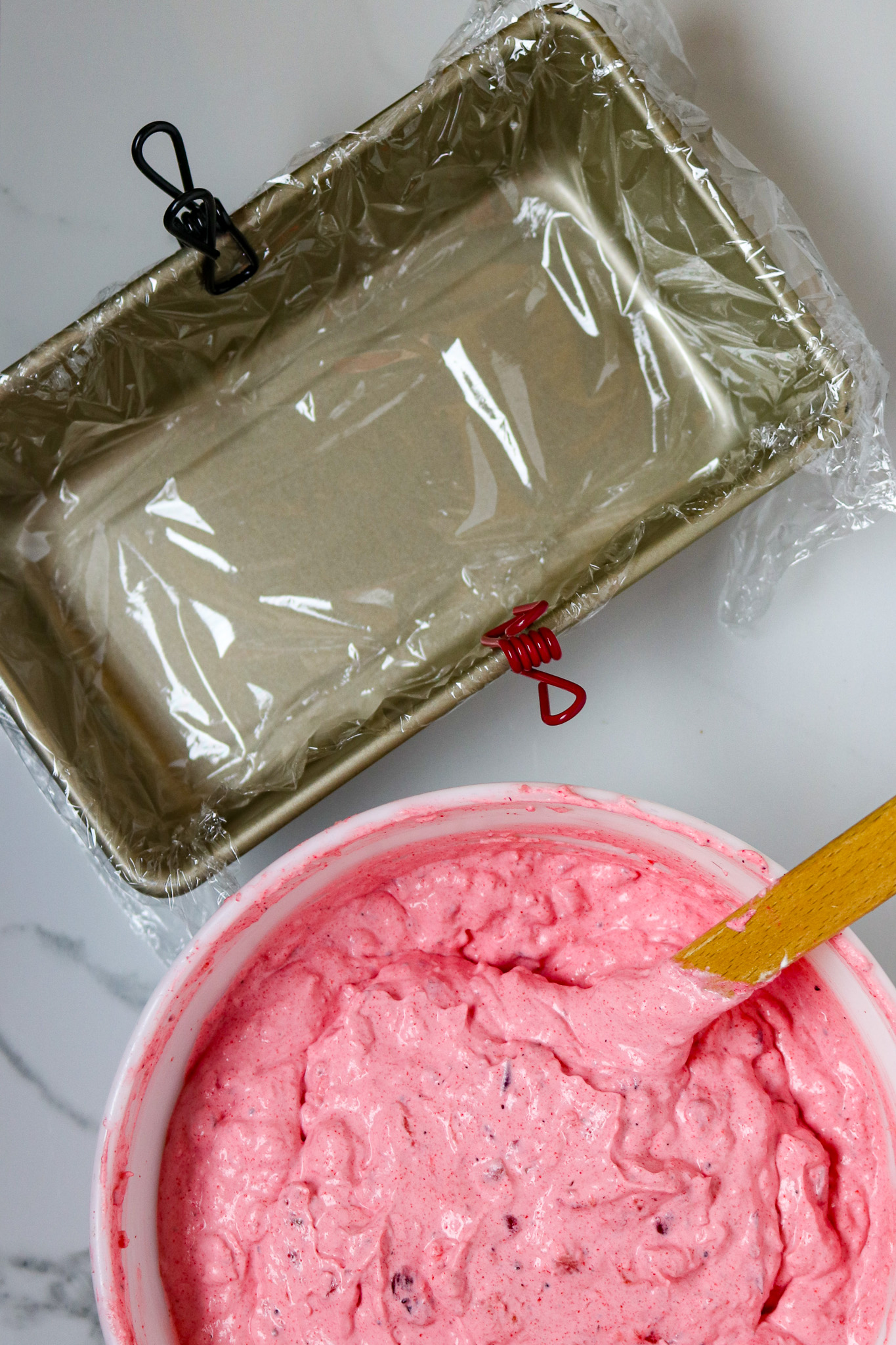 A bread loaf pan lined with saran wrap next to a bowl of pink whipped dessert.
