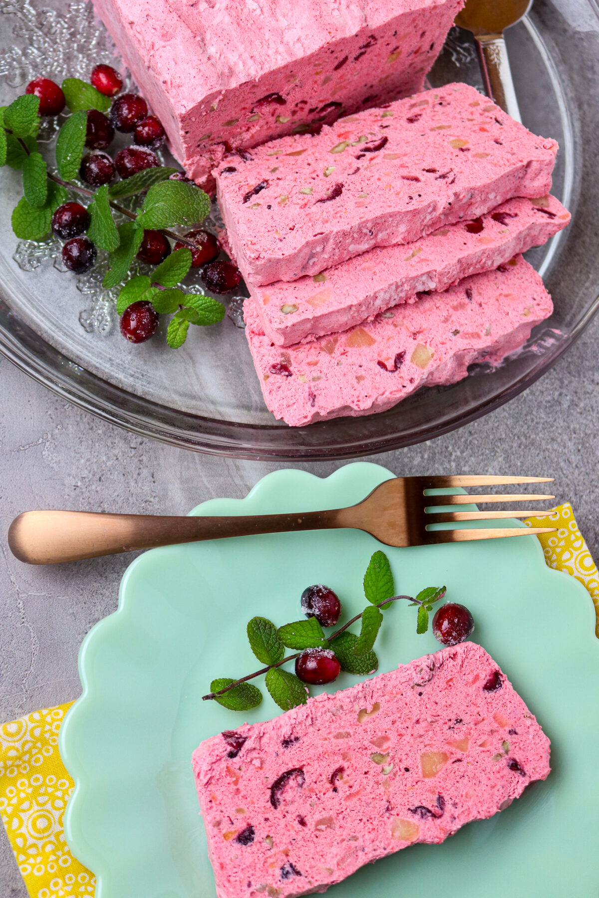 A loaf of frozen cranberry salad sliced with fresh cranberries and mint leaves with a fork and a sliced serving on a green scalloped edge plate.