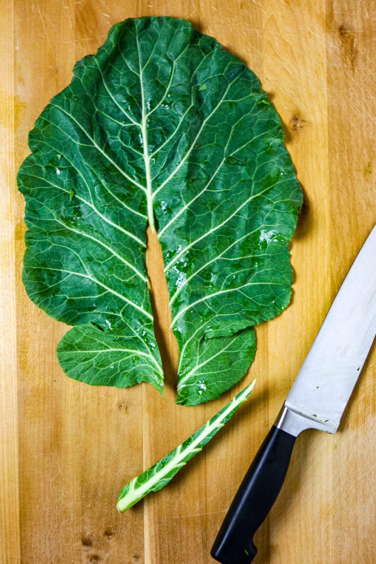 A leaf of collard green with stem sliced out of it on a wooden board,