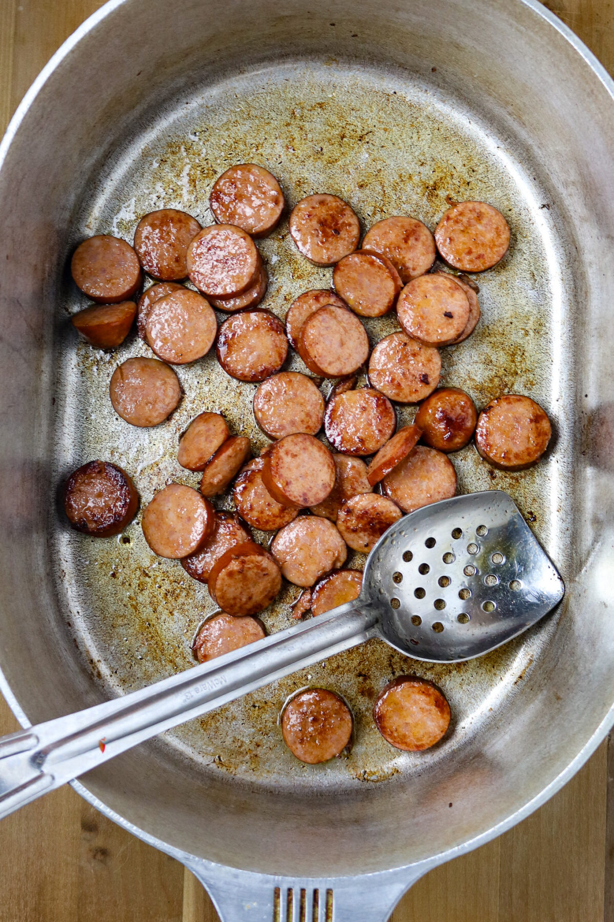 Slices of smoked sausage browned in a large silver pot with a slotted spoon in it.