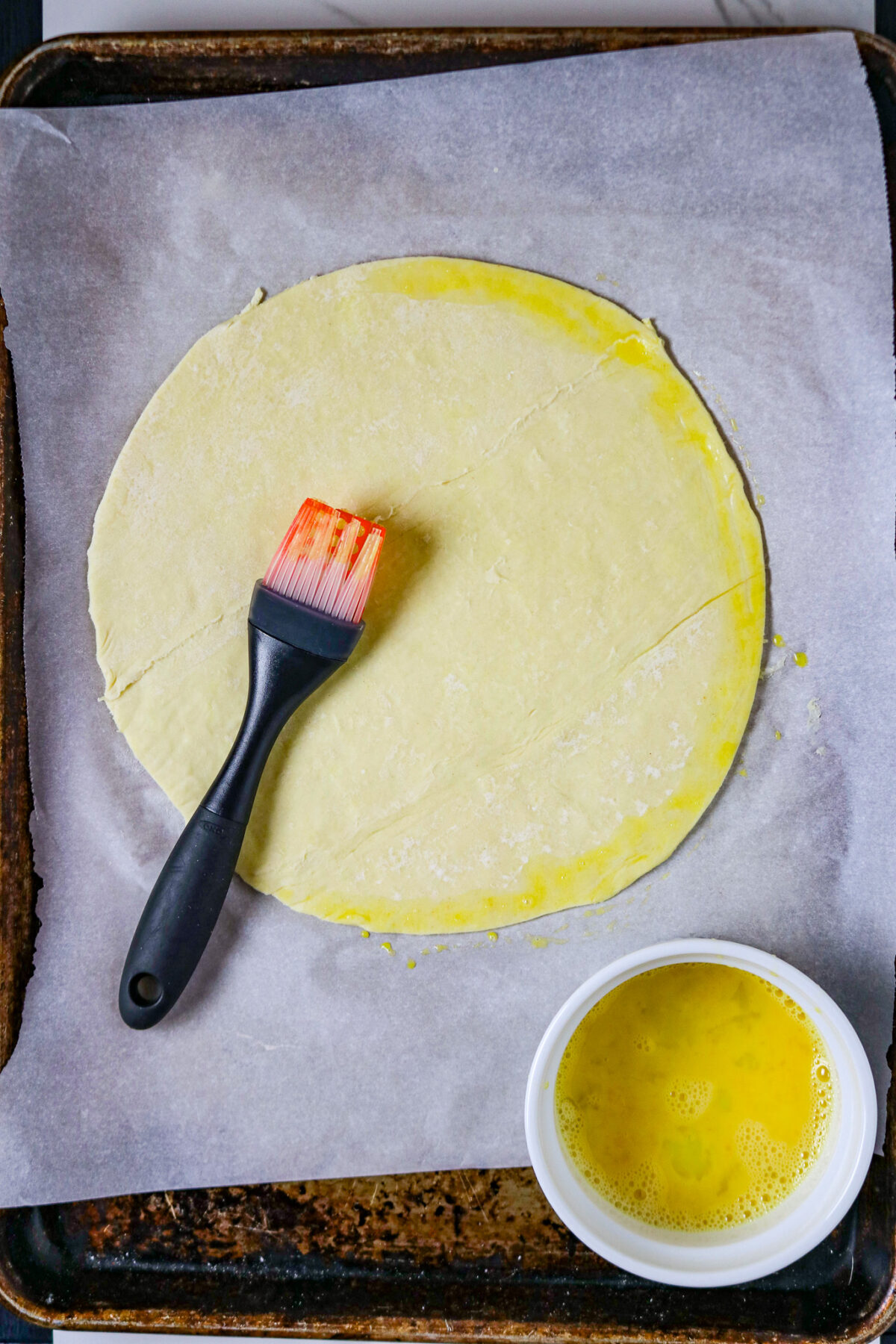 A round puff pastry dough on a parchment paper with a pastry brush laying on top.
