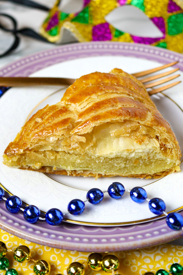 A slice of Galette Des Rois on a white plate with a brass fork and a string of purple Mardi Gras beads.