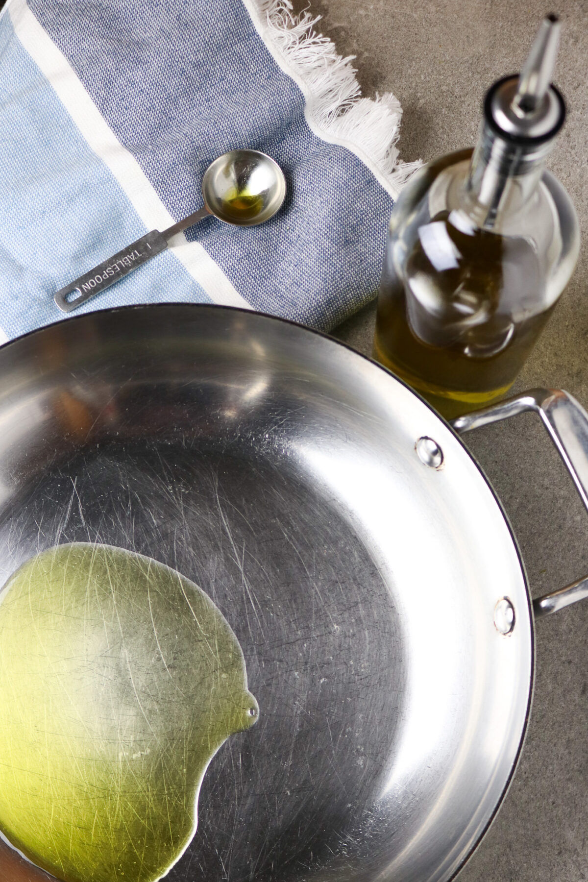 A large stainless skillet with a small amount of oil in it and a measuring spoon an a bottle of oil next to it with a blue dish towel.