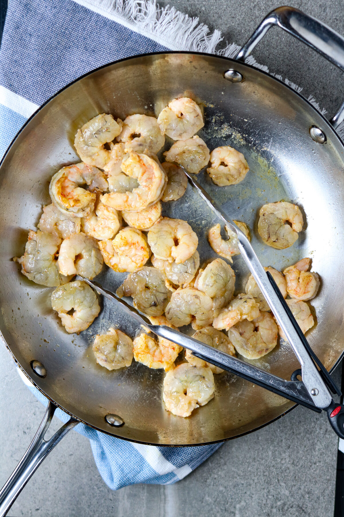 A large stainless skillet with shrimp and cooking tongs in it.