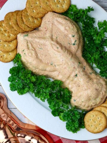 A white plate with a congealed shrimp dip in the shape of a crawfish surrounded with fresh parsley and crackers next to a copper colored crawfish mold.