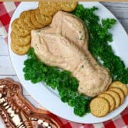 A white plate with a congealed shrimp dip in the shape of a crawfish surrounded with fresh parsley and crackers next to a copper colored crawfish mold.