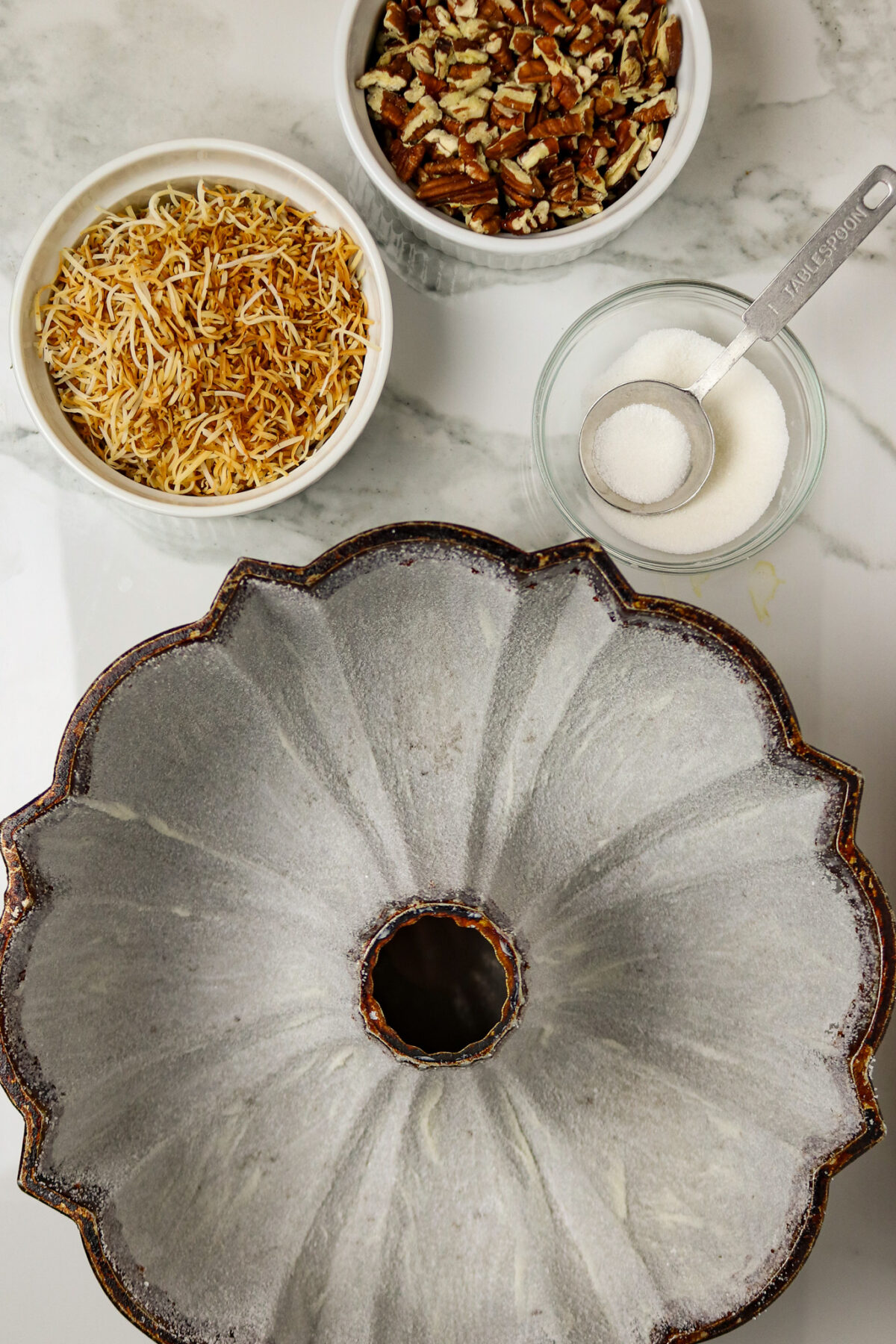 A prepared bundt cake pan coated with sugar and bowls of sugar, coconut, and pecans beside it.