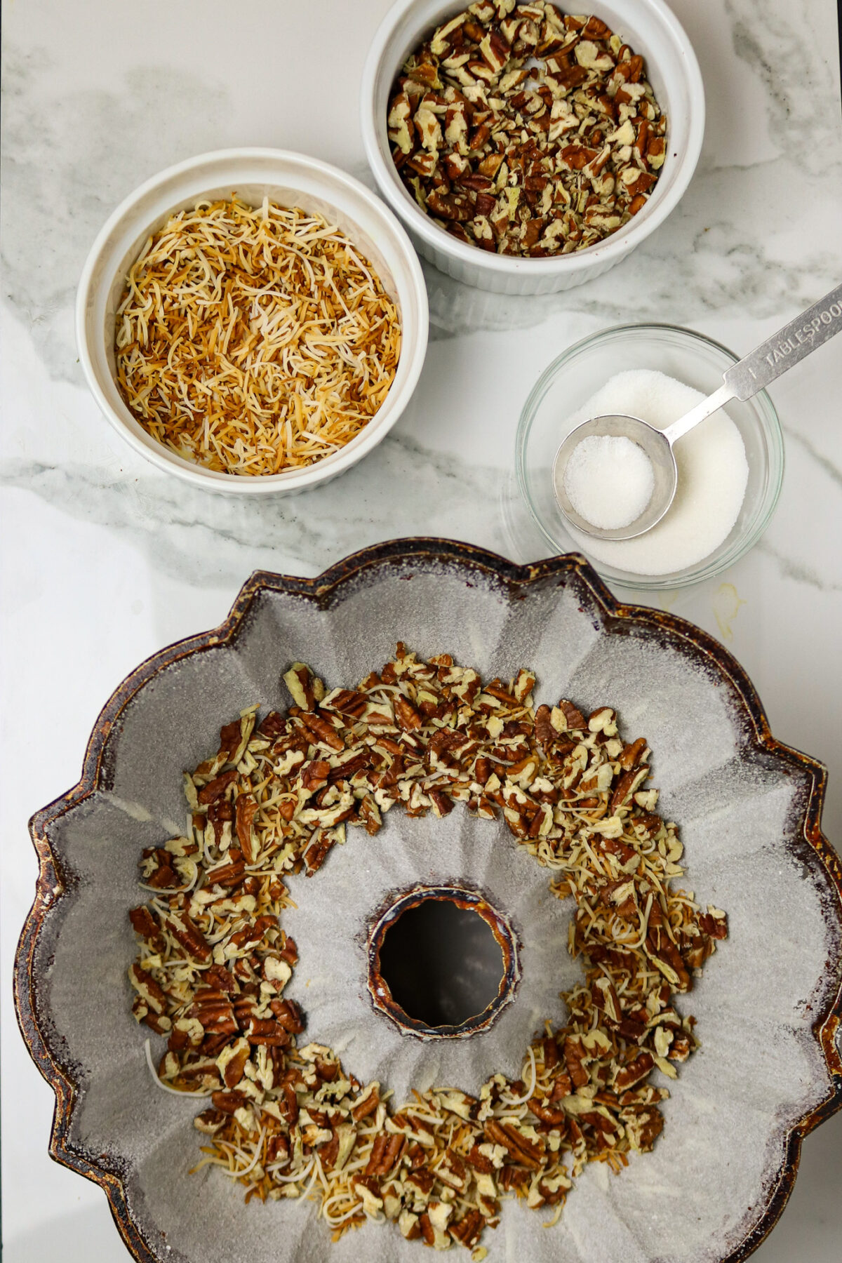 A prepared bundt cake pan with nuts and coconut on the bottom and bowls of sugar, coconut, and pecans beside it.