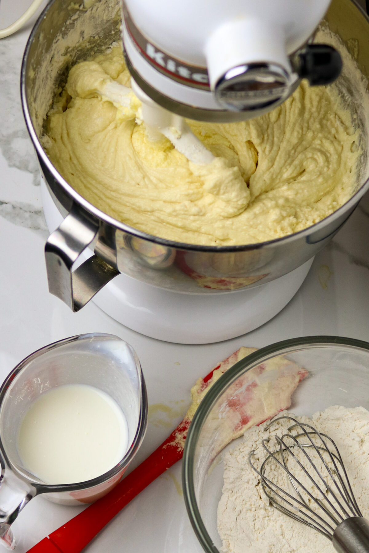 A stand mixer with batter in the bowl and a cup of buttermilk and a bowl of dry ingredients next to it.