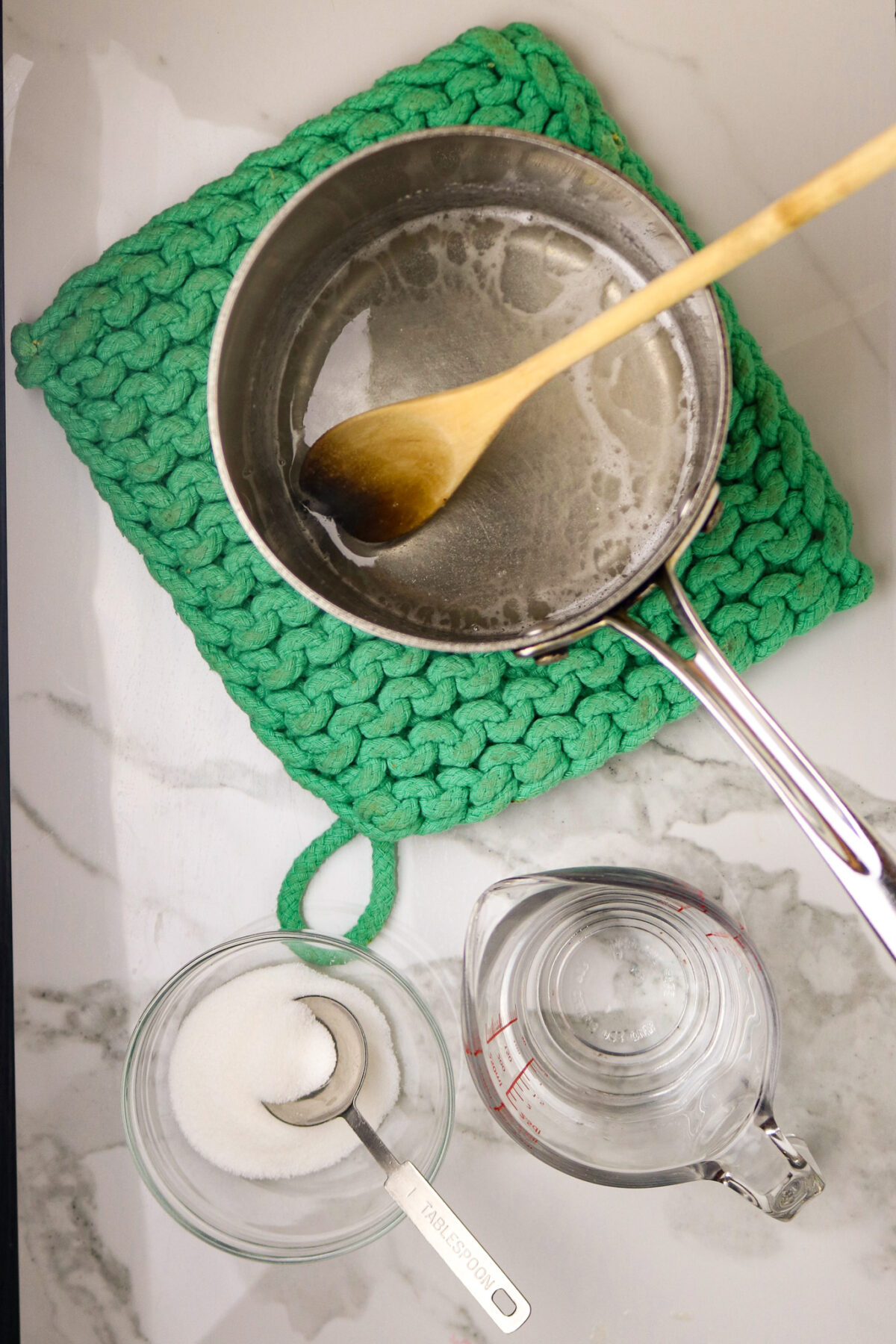 A saucepan with a wooden spoon stirring simple syrup and a cup of sugar and water net to it.