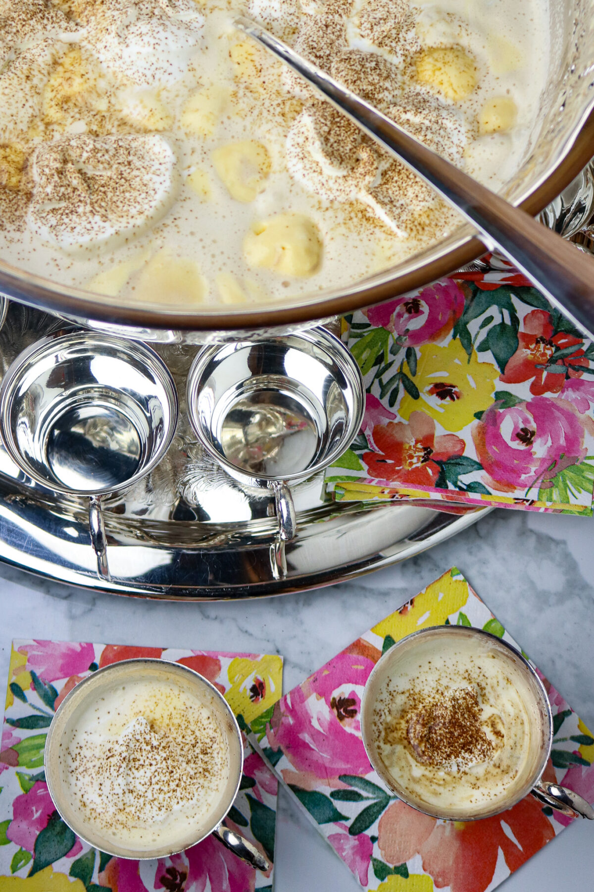 A large. silver punch bowl filled with punch on a silver platter with silver punch cups.