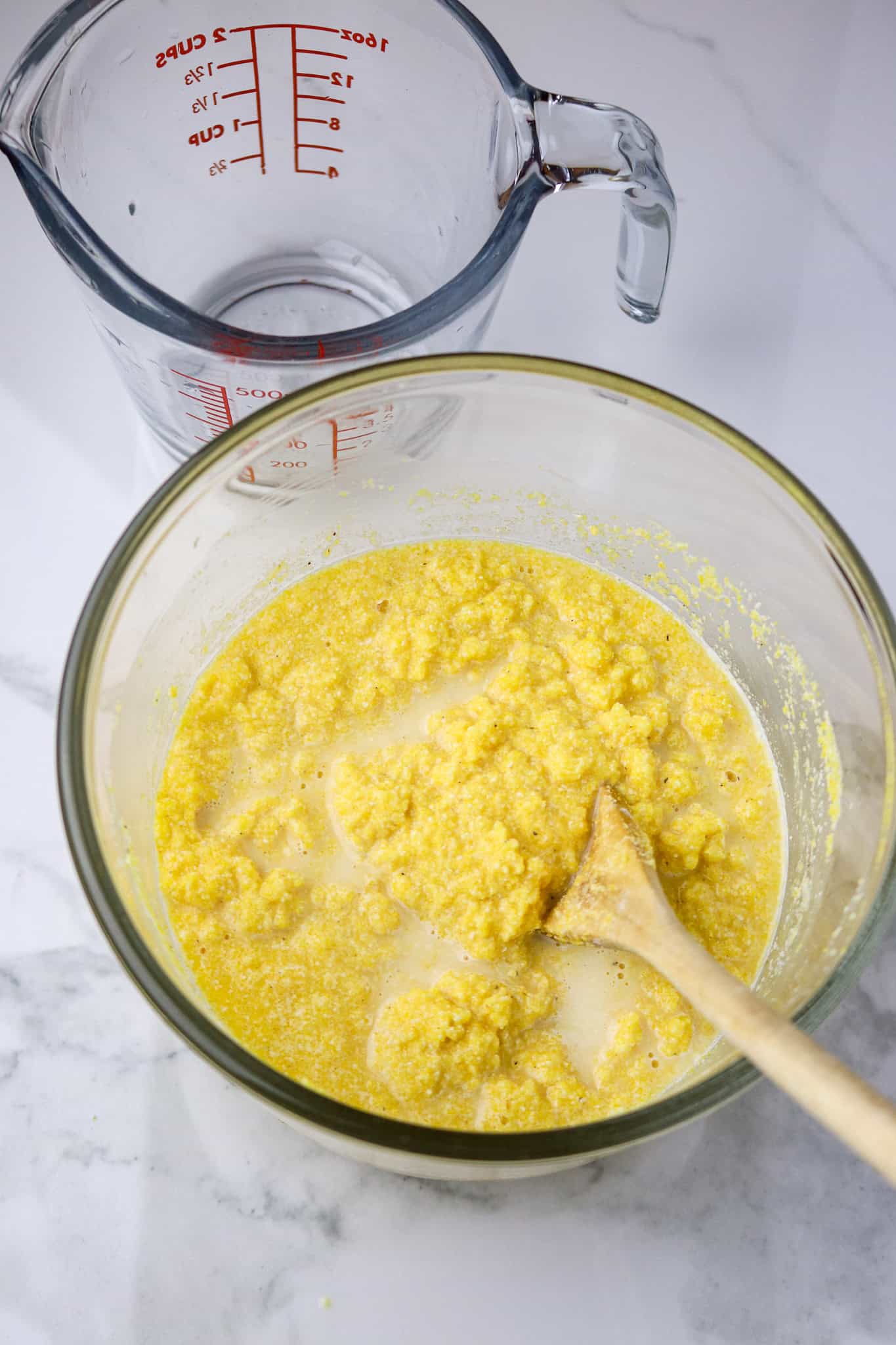 A glass bowl with corn meal mush in is and a wooden spoon for couche couche recipe.