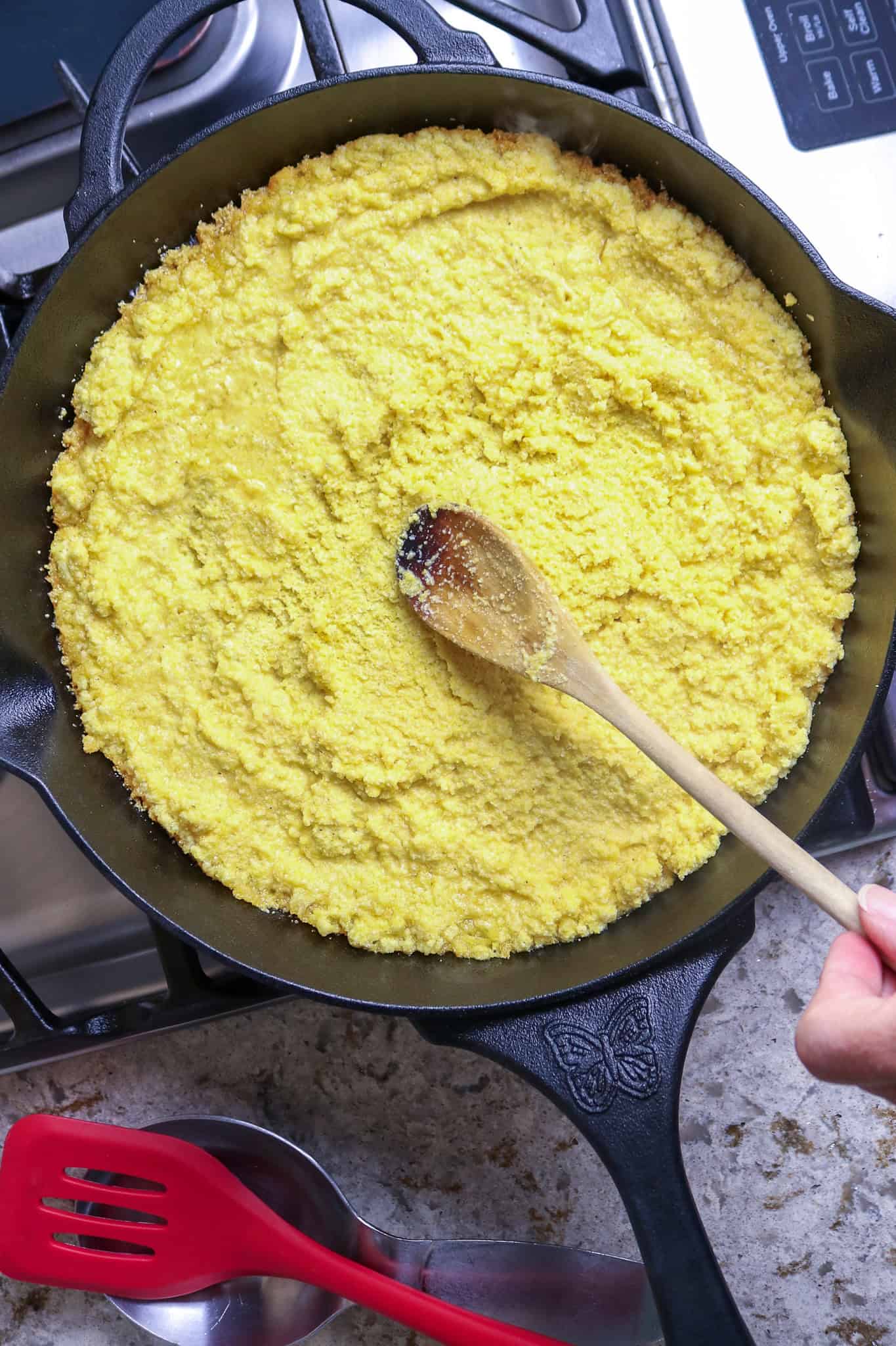 A wooden spoon spreading corn meal mush into a black iron skillet for couche couche recipe.