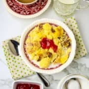 A white rimmed bowl on a green polk a dotted napkin with a spoon filled with couche couche recipe and milk.