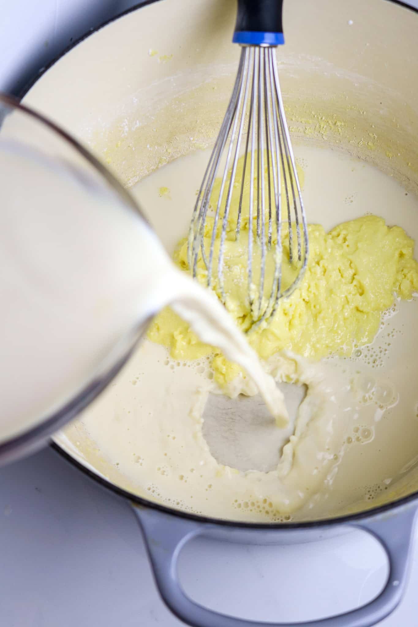 A glass meaturing cup pouing milk into a pot with a whisk in it.