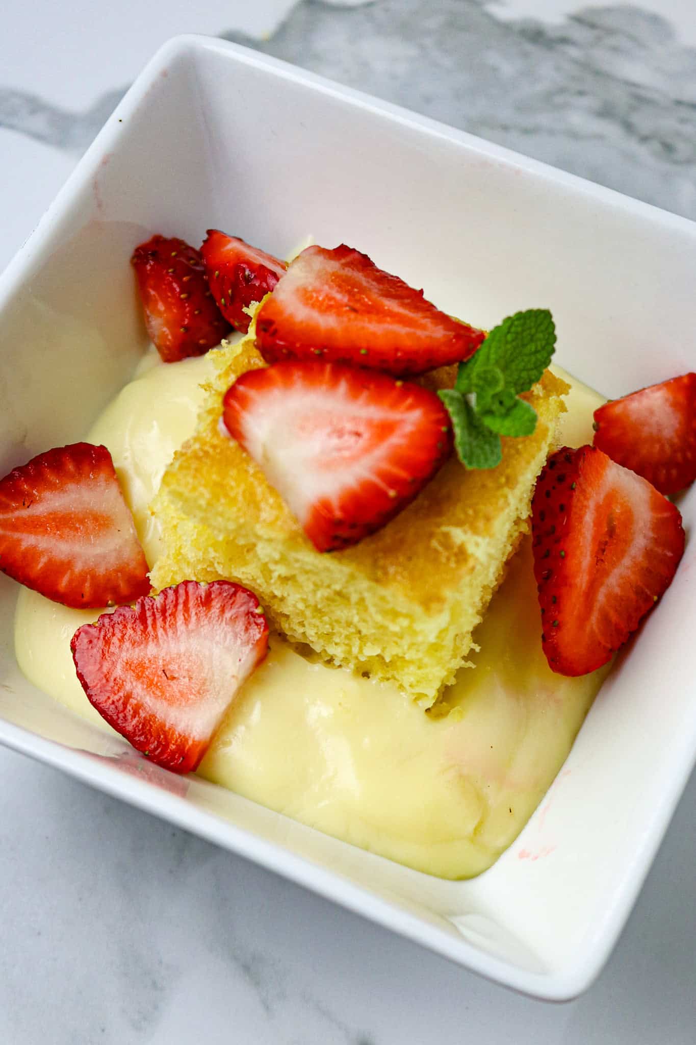 A white square dish with bouille custard, a square of cake, and berries with mint leaves on top.