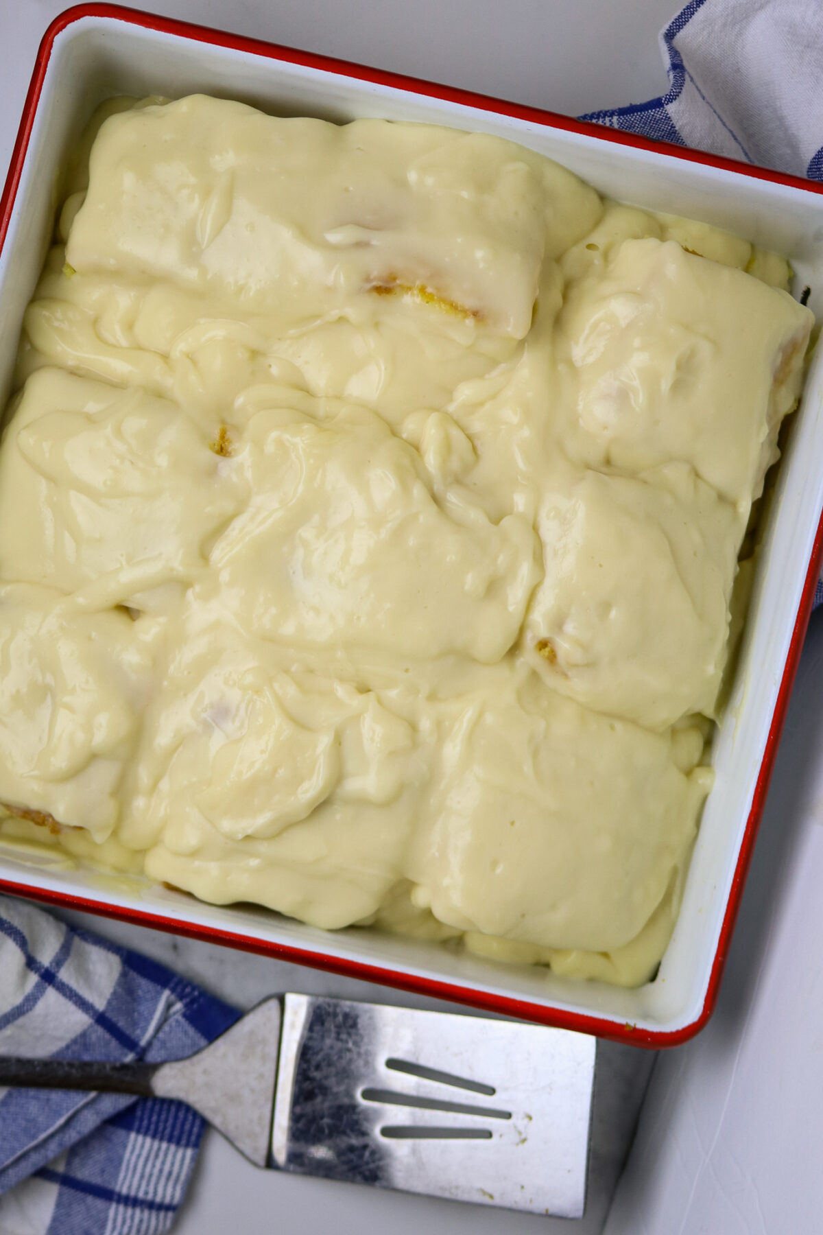 A square pan with a red trim filled with cake squared and covered with custard.