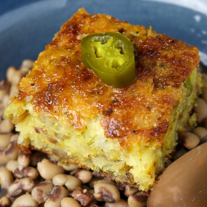 A square piece of cornbread on a bowl full of black eye peas and a slice of jalapeno pepper on top of the bread..