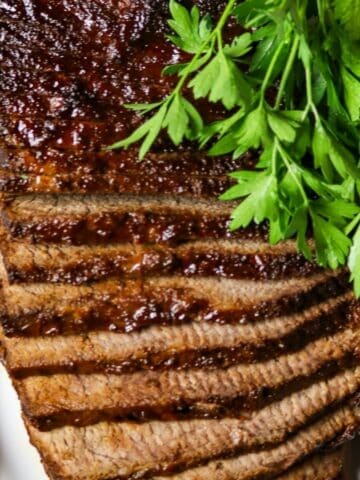 A sliced, cooked beef brisket on a white plate with a bunch of fresh parsley.