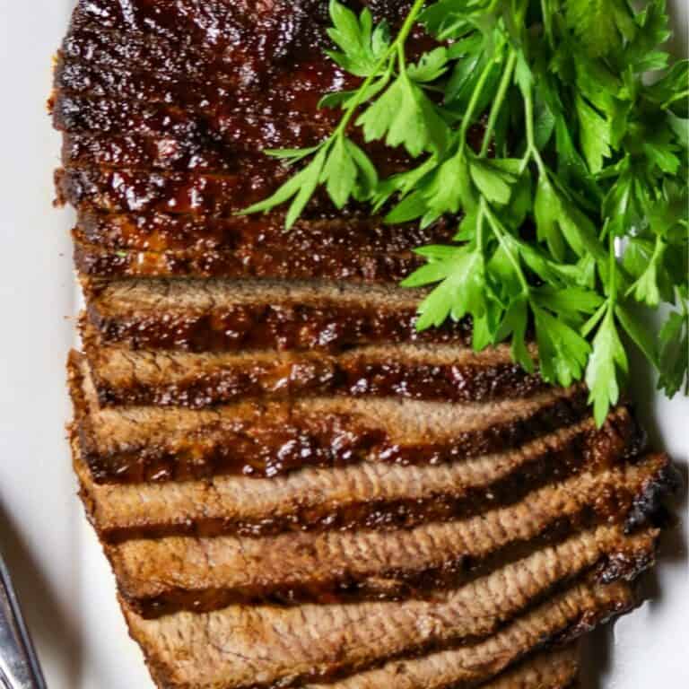 A sliced, cooked beef brisket on a white plate with a bunch of fresh parsley.