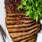A sliced, cooked beef brisket on a white plate with a bunch of fresh parsley.