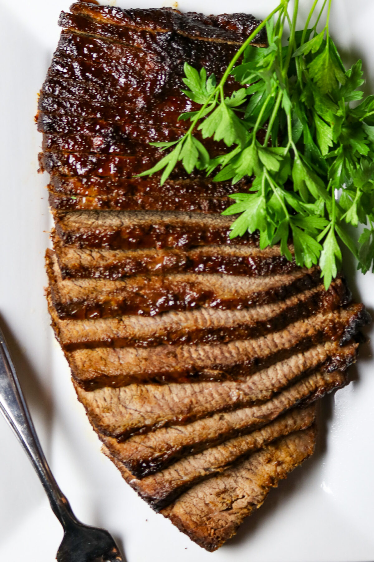 A sliced, cooked beef brisket on a white plate with a bunch of fresh parsley.