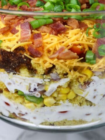 A large glass bowl of layers of salad ingredients of peppers, corn, onion, bacon, cheese, and cornbread.