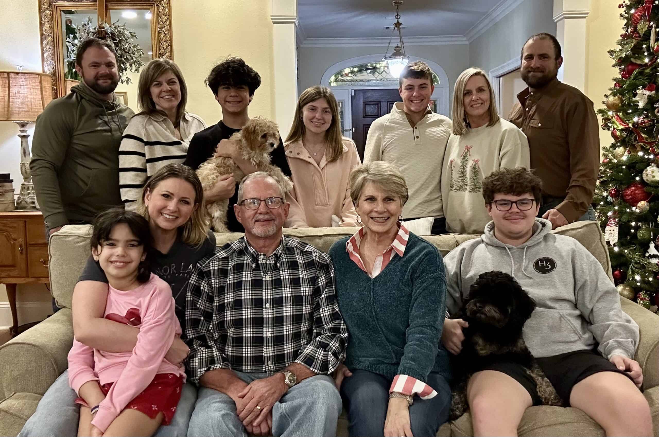 A family photo of twelve people and two dogs sitting on a green couch and standing behind it with a Christmas tree on the side.