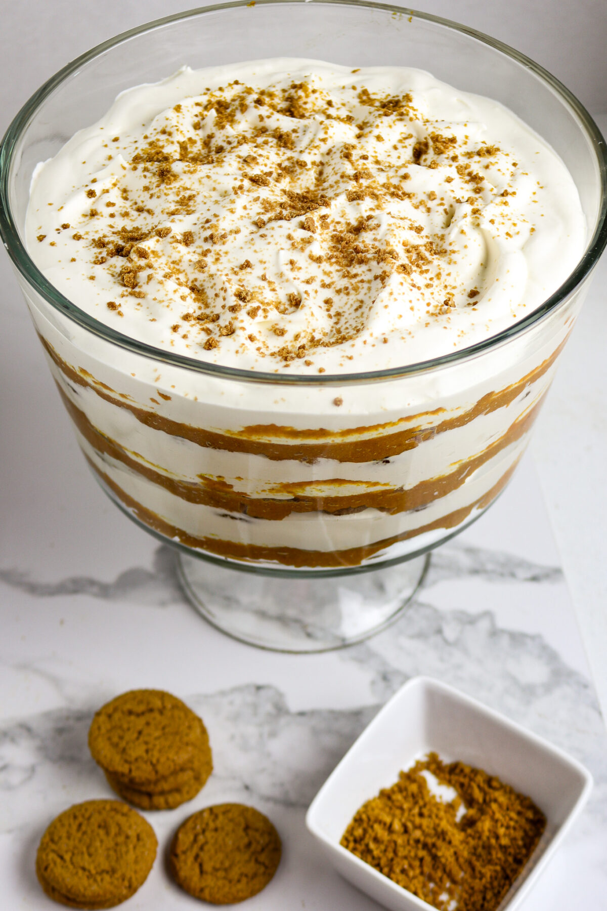 A footed glass dish with 6 layers of cookies, pumpkin, and whipped cream with cookie sprinkles on top for a ginger snaps pumpkin trifle dessert.