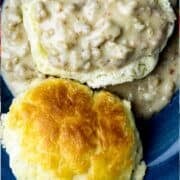 A black-iron skillet of biscuits next to a blue bowl with a biscuit and homemade sausage gravy poured on top.