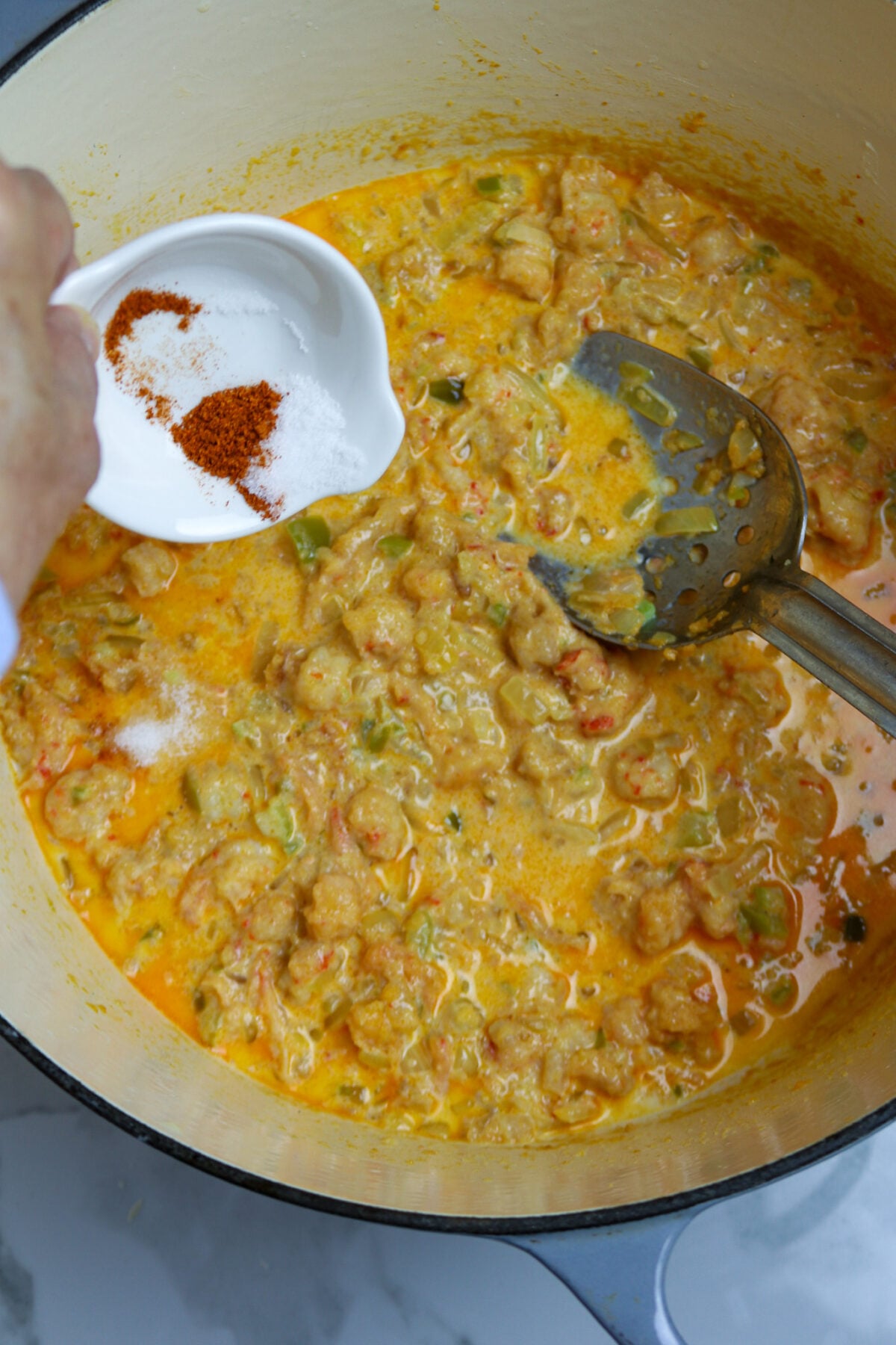 A large pot with crawfish in a cheese sauce, a small white bowl of seasonings poured over the sauce, and a silver spoon in the pot.