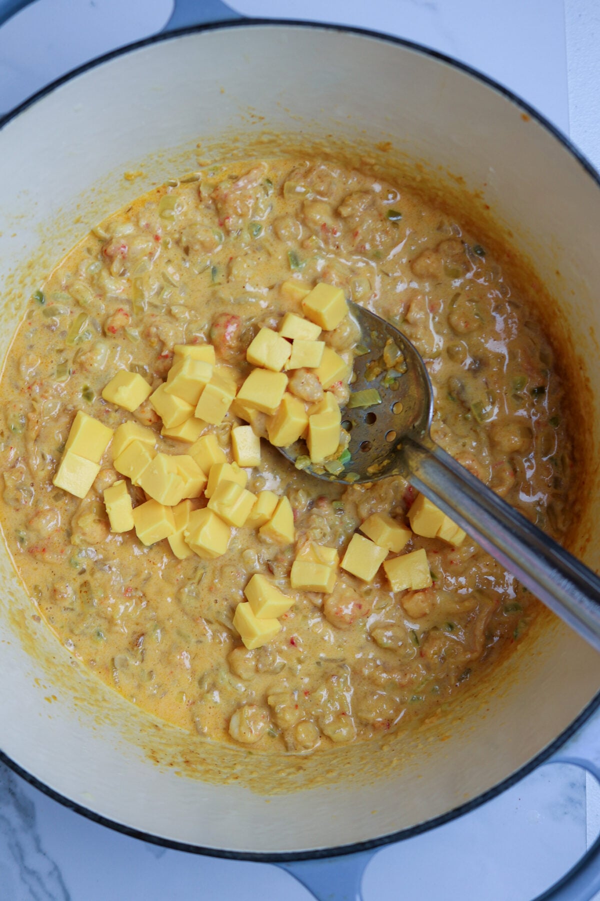 A large pot with crawfish in a sauce with cheese cubes and a silver spoon in the pot.