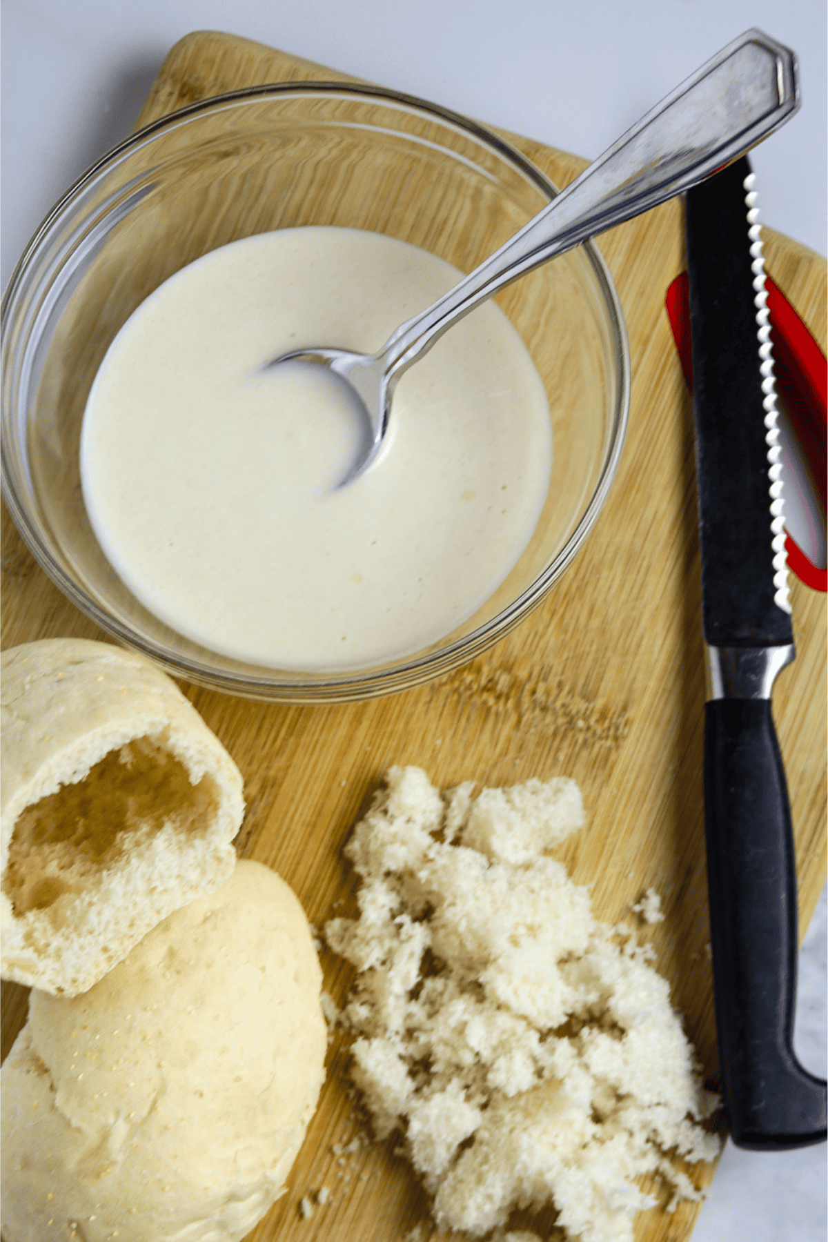 A hallowed out small French bread roll on a wooden cutting board with a bowl of white sauce and a serrated knife.