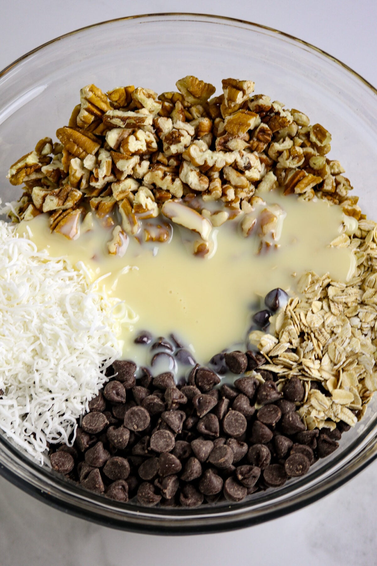 A glass bowl separately showing chopped pecans, oatmeal, chocolate chips, coconut, and condensed milk before mixed together.