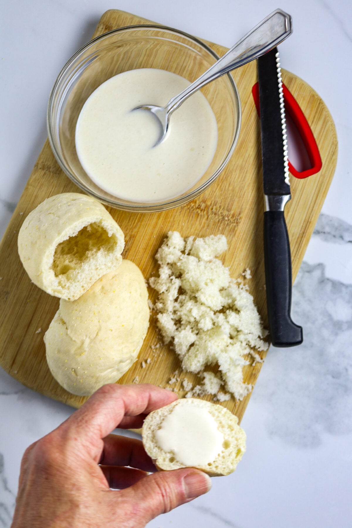 A wooden cutting board with a pistolette bun cut in two with bread crumbs taken out and a bowl of white flour paste, and a knife.