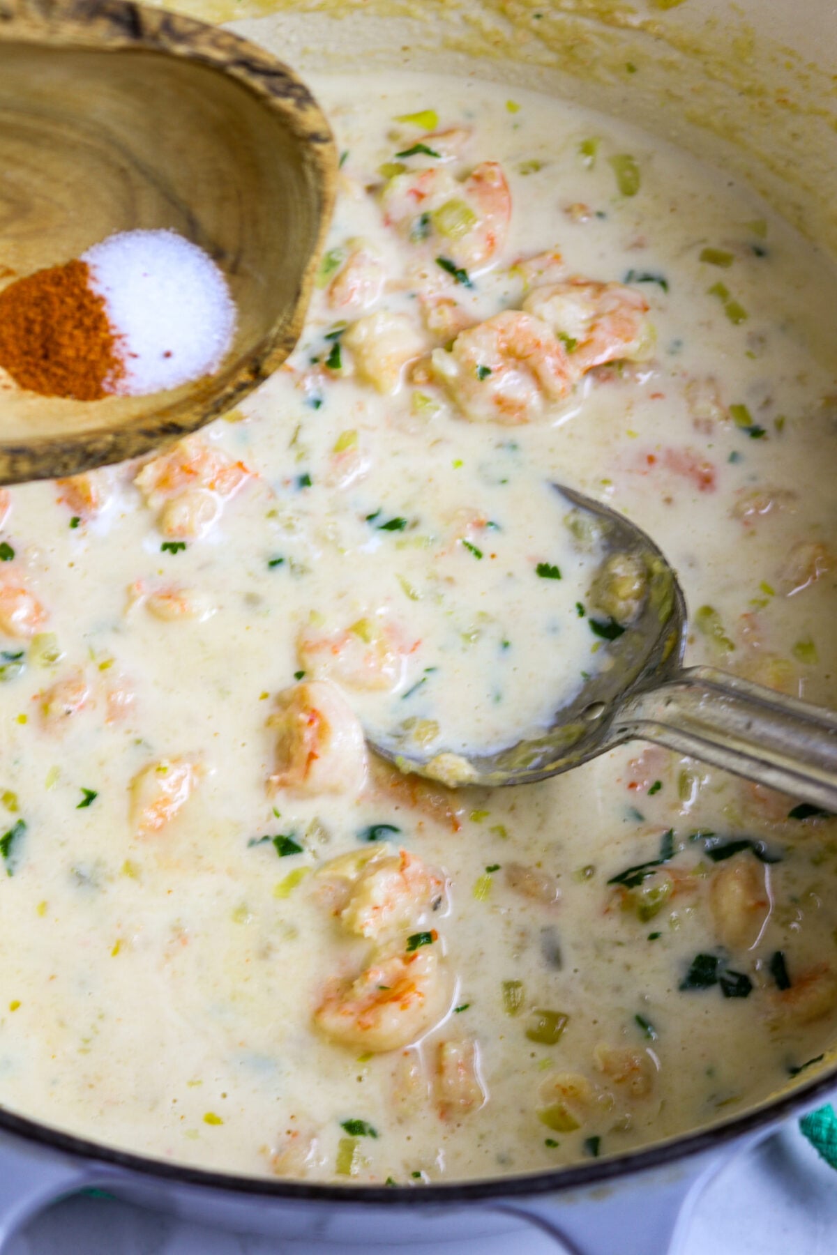 A wooden bowl of spices over a pot of reamy shrimp sauce with a silver spoon in it.