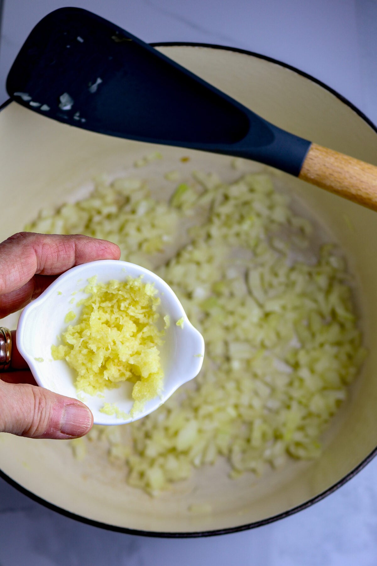 A small bowl of minced garlic over a pot of chopped onion with a spoon on the pot's edge.